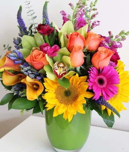  arrangement features sunflowers, gerbera daisies, roses, orchids, calla lilies & more. Designed in a beautiful green glass vase. This bright & cheerful floral arrangement is sure to bring a smile! 