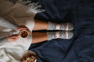 Woman in warm socks with cup of hot drink relaxing on knitted blanket, top view